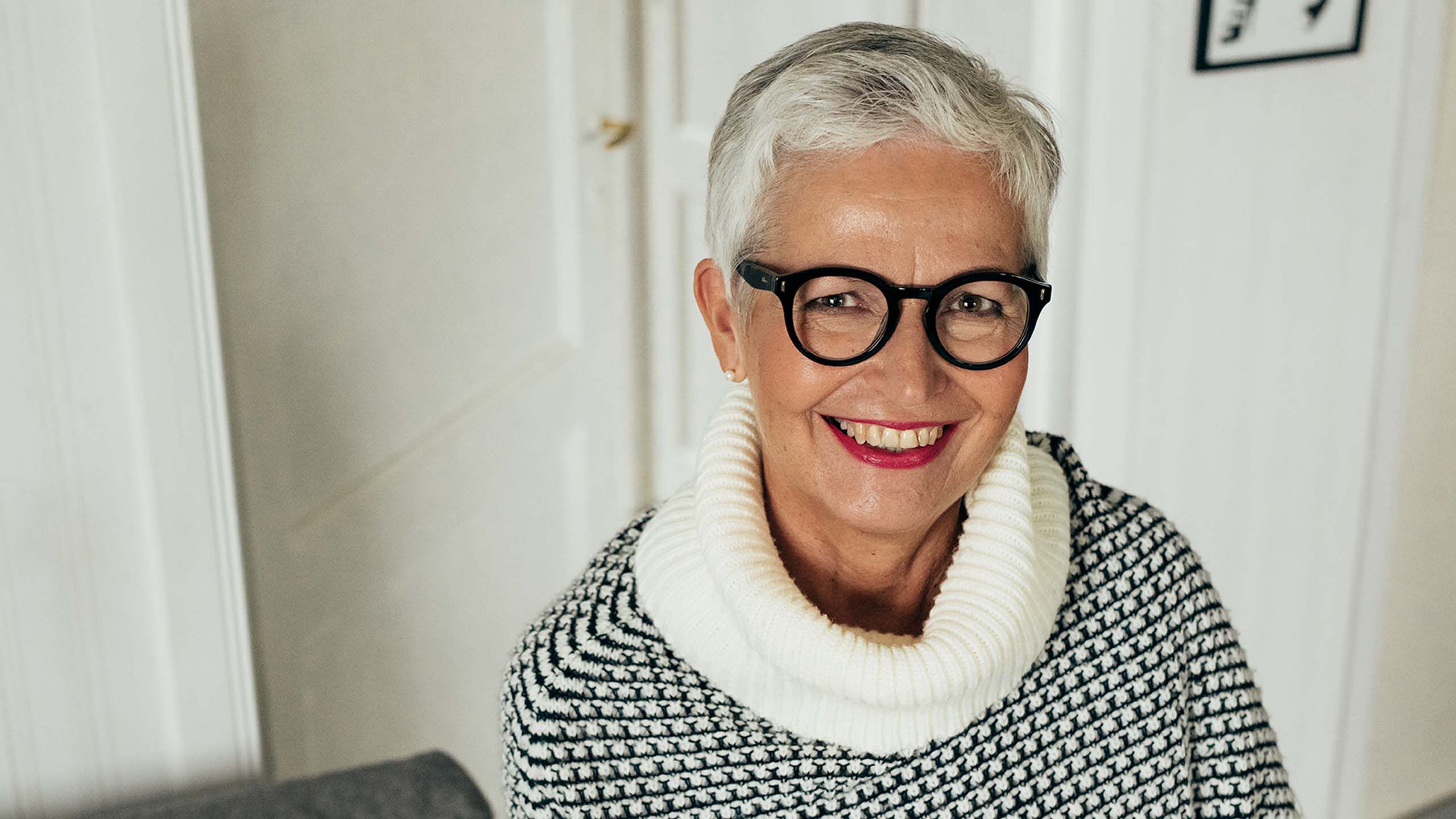 Rouva istuu olohuoneen sohvalla, yll&auml;&auml;n villapaita ja mustat silm&auml;lasit